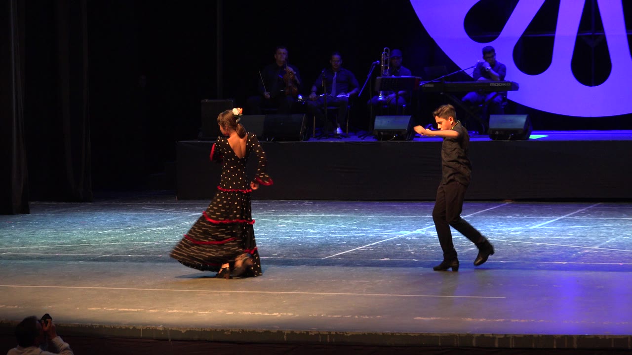 The two Cabo San Lucas performers remain separated across the stage, with the dress hem spread near the floor and the boy extending his arms under blue-purple stage light.