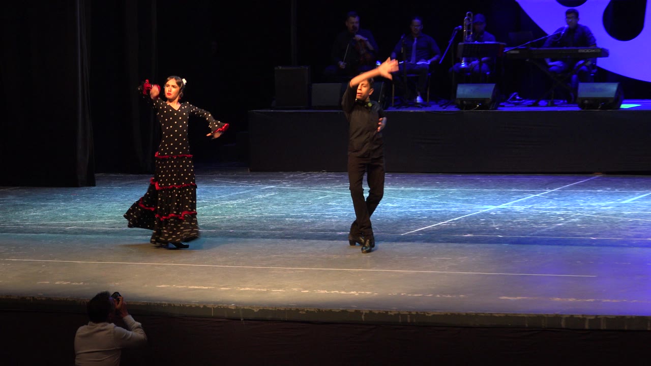 At 0:00 in Cabo San Lucas, Mexico, a woman in a black flamenco dress and a man in black hold raised arm poses on a stage while musicians sit in blue backlight behind them.