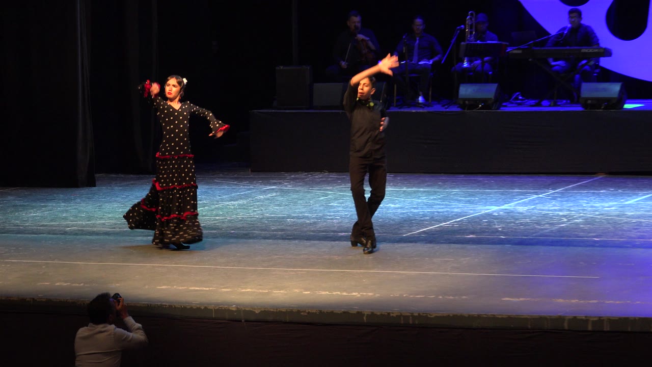 At 0:01 in Cabo San Lucas, Mexico, the dancers remain in a wide stage view, with the woman holding her dress and the man lifting one arm near the blue-lit band platform.