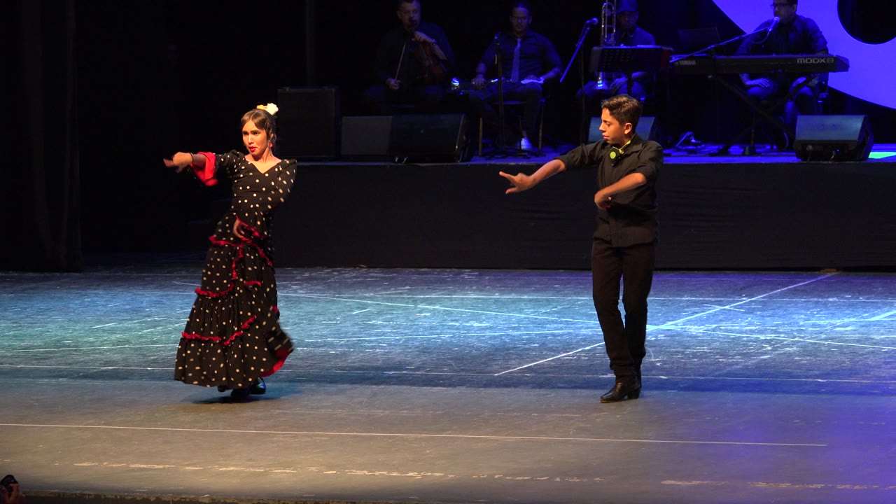 In Cabo San Lucas, Mexico, a woman in a black polka-dot dress and a male partner in black extend their arms on a blue-lit stage with musicians seated in the background.