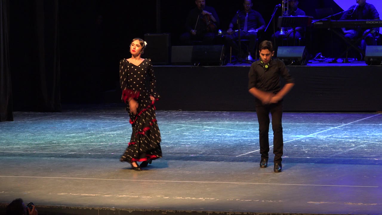Two dancers stand on a theater stage with musicians behind them.