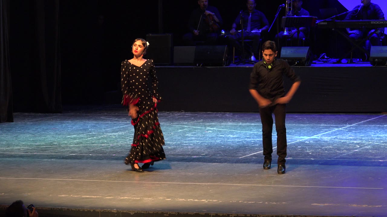 The dancers hold a measured starting pose on the Cabo San Lucas stage, with the woman looking forward and the man angled downward while musicians remain dimly lit behind them.