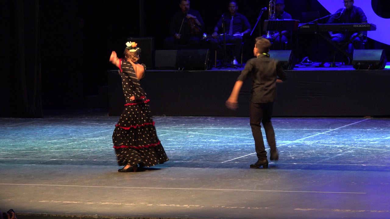In Cabo San Lucas, Mexico, the woman lifts one arm while the man steps away from her across the stage, with musicians dimly visible behind them.