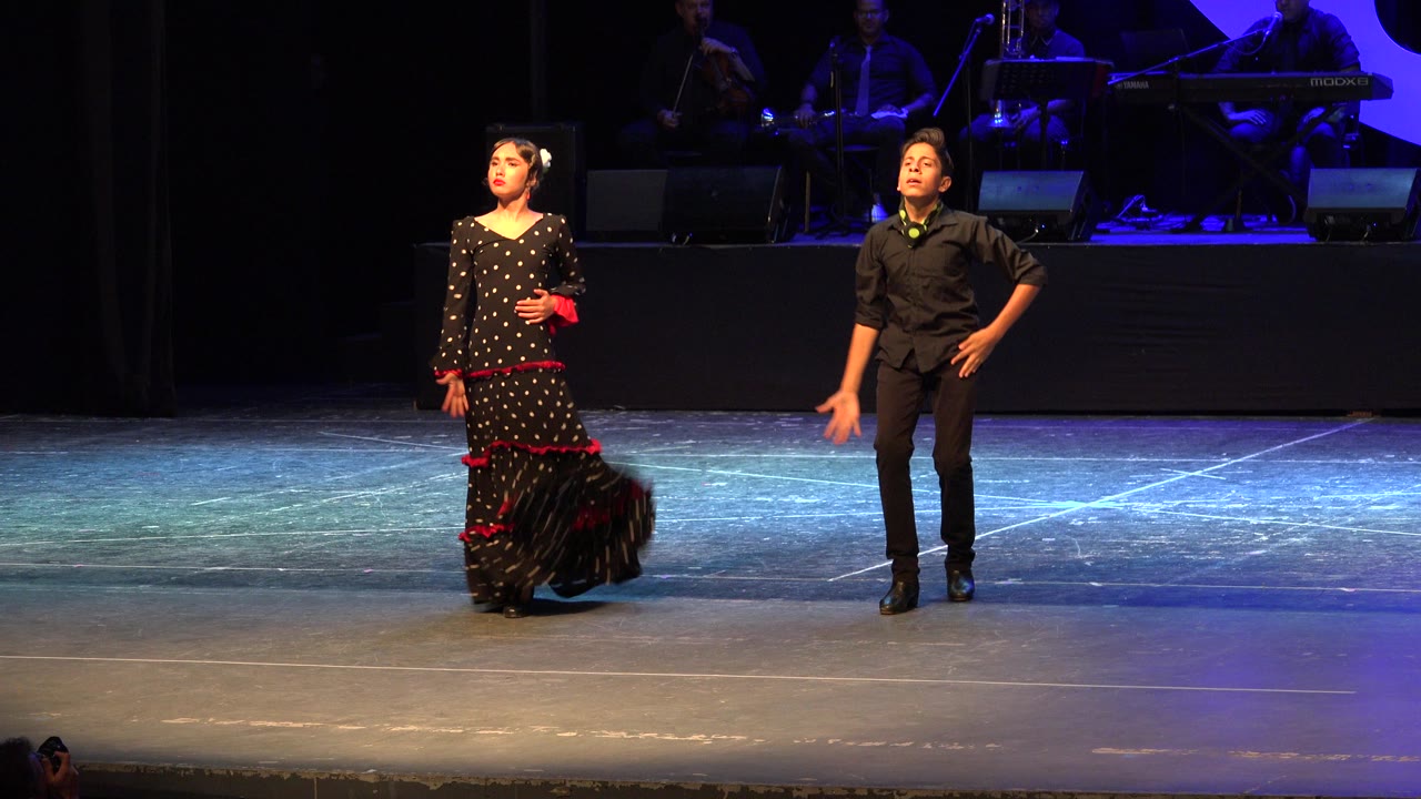Two young flamenco dancers stand on a lit stage with musicians in the background.