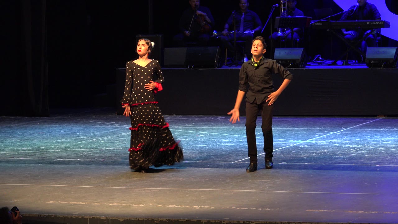 The young dancers remain in a wide stage composition in Cabo San Lucas, with the girl holding one hand near her waist and the boy standing with one hand on his hip.
