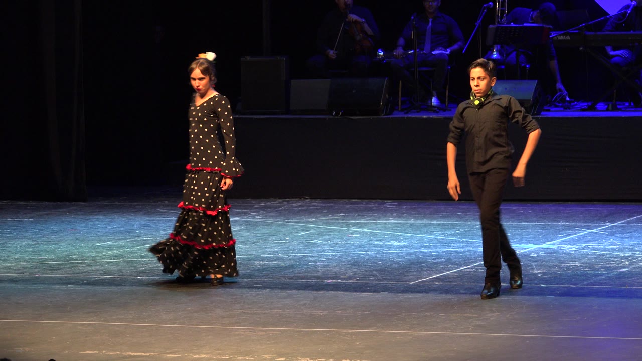 Two young dancers stand on a dark stage under blue lighting.