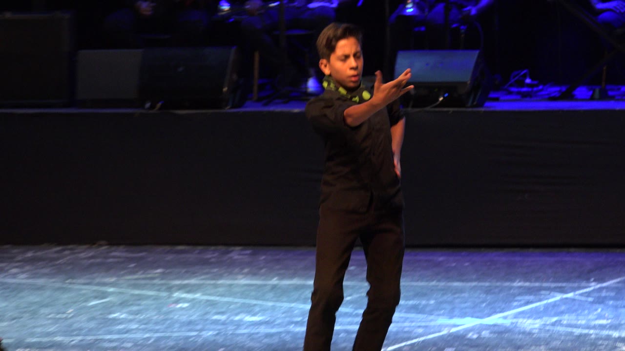 Young dancer in black reaches one arm forward on a dark blue-lit stage.