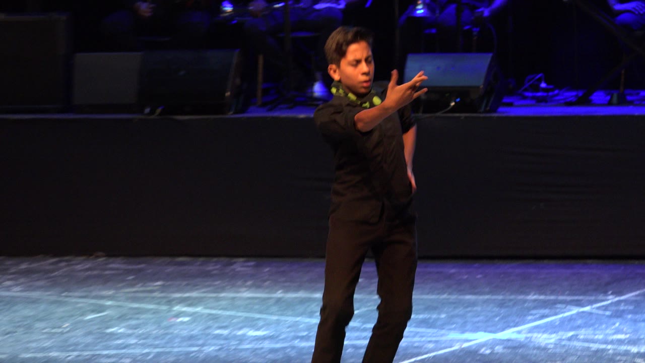 Young dancer in black holds a forward gesture under blue-purple stage light in Cabo San Lucas, Mexico, with dark performance equipment in the background.