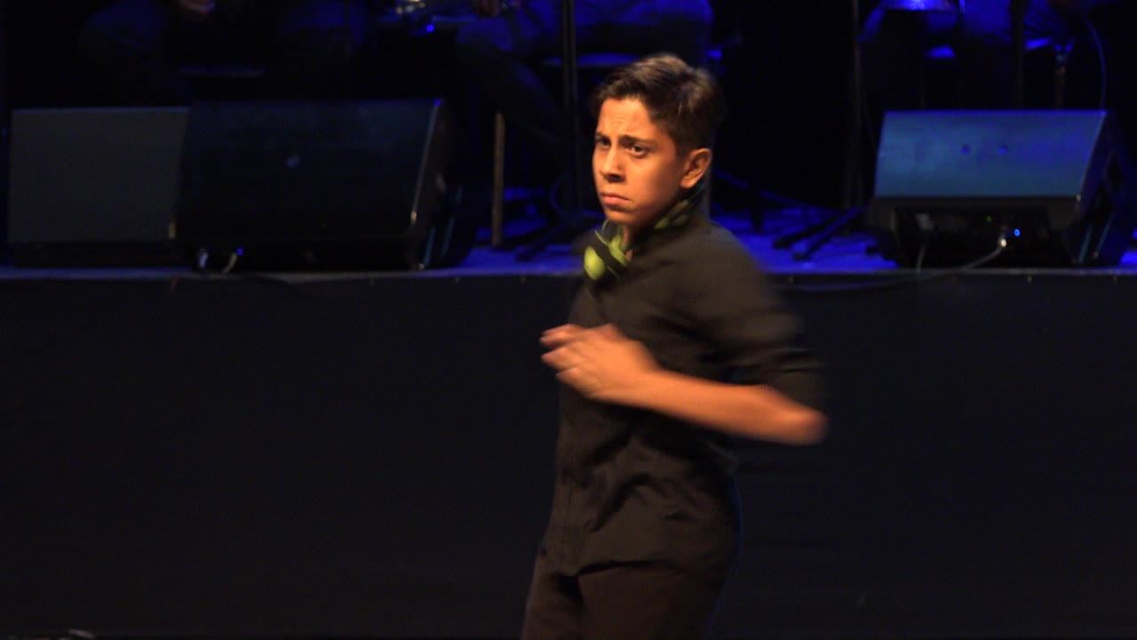 Young dancer in black shirt with green scarf on dark stage.