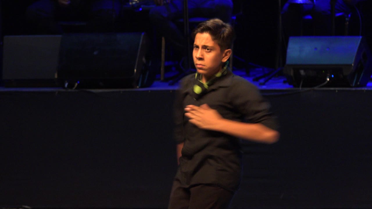 Young dancer in black clothing keeps one hand near his chest while standing against a dark stage background with blue-lit monitors in Cabo San Lucas, Mexico.