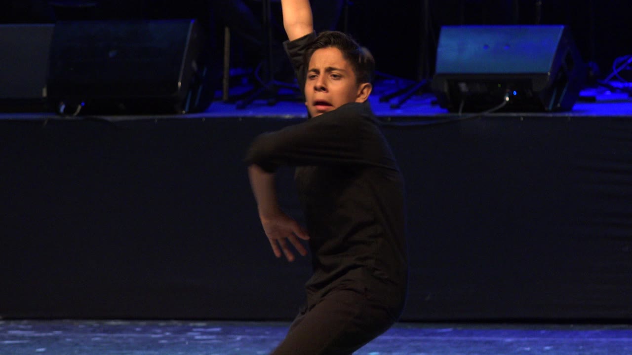 Young boy in black raises one arm while dancing on a dark blue-lit stage in Cabo San Lucas, Mexico.