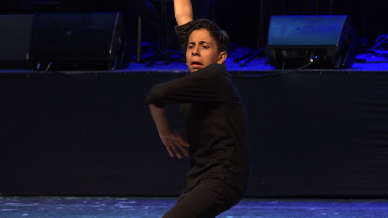 Young boy in black keeps one arm lifted and the other bent across his body on a dim stage in Cabo San Lucas, Mexico.