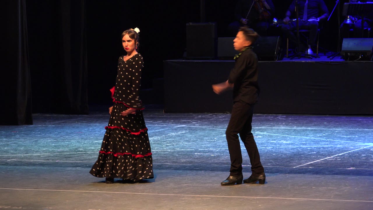 The youth dancers continue their stage routine in Cabo San Lucas, Mexico, with the girl holding her skirt area and the boy stepping through a blurred arm movement near the back line.