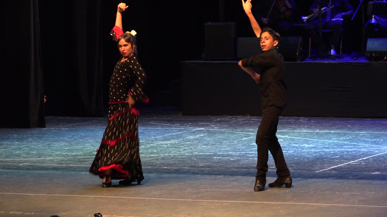 Two flamenco dancers on a dark indoor stage with arms raised.