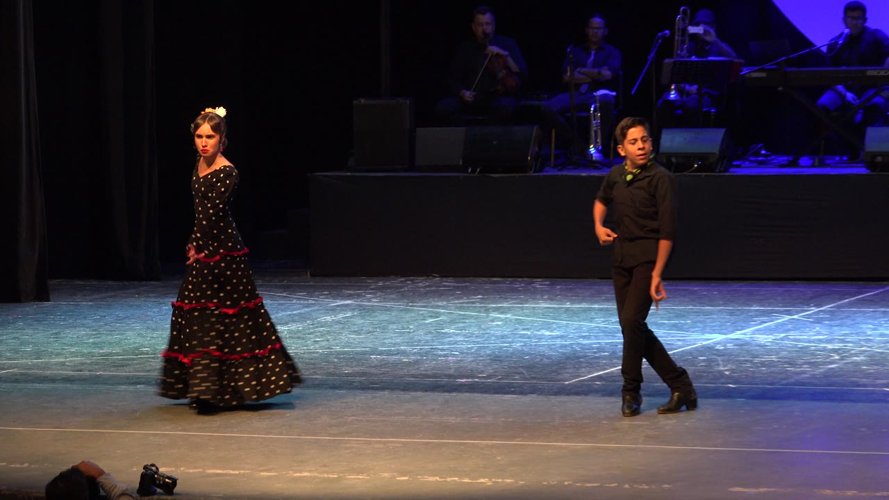 At 0:01, the dancers remain separated across the Cabo San Lucas stage as the girl faces forward in her black dress and the boy stands angled in black clothing near the center-right.