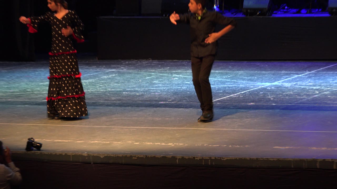 The girl in a black dress with red trim and the boy in black continue their dance on the Cabo San Lucas stage, with floor markings visible under colored light.