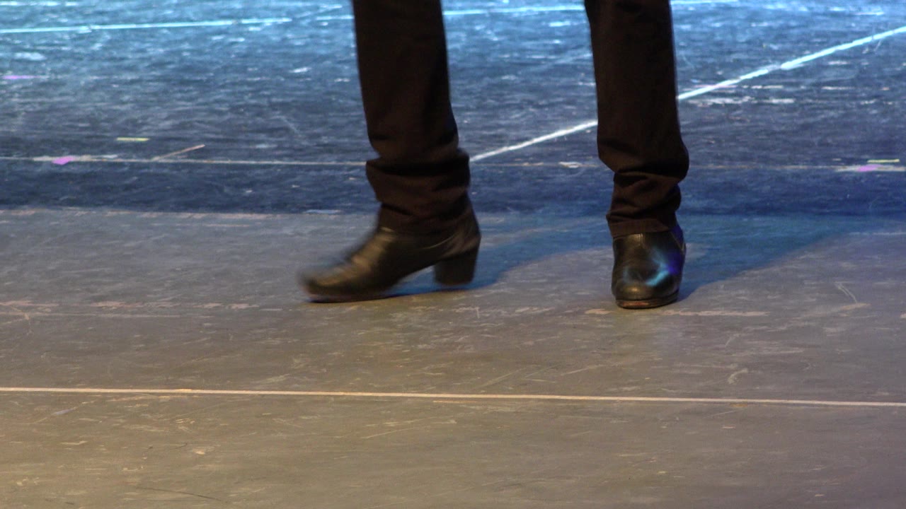The dancer's black shoes are set apart on the stage floor, with cool blue light and white floor lines visible behind them in Cabo San Lucas, Mexico.