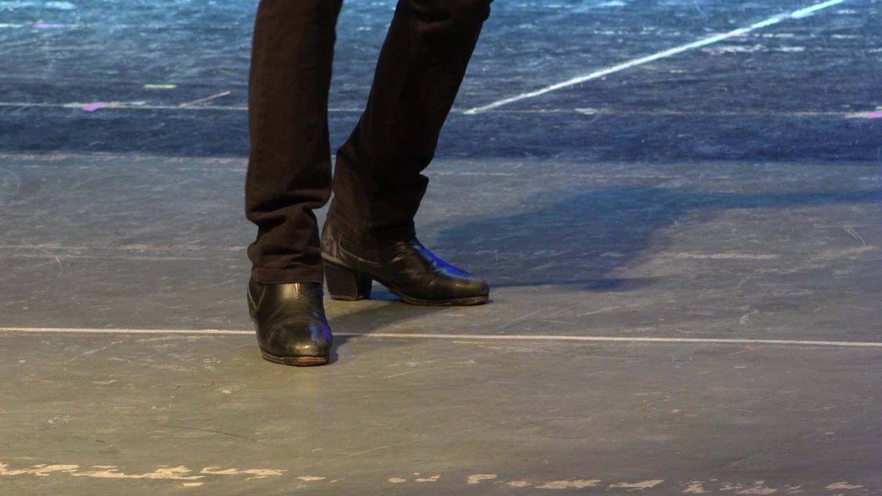 Cropped lower legs in black pants and black shoes on a stage floor.