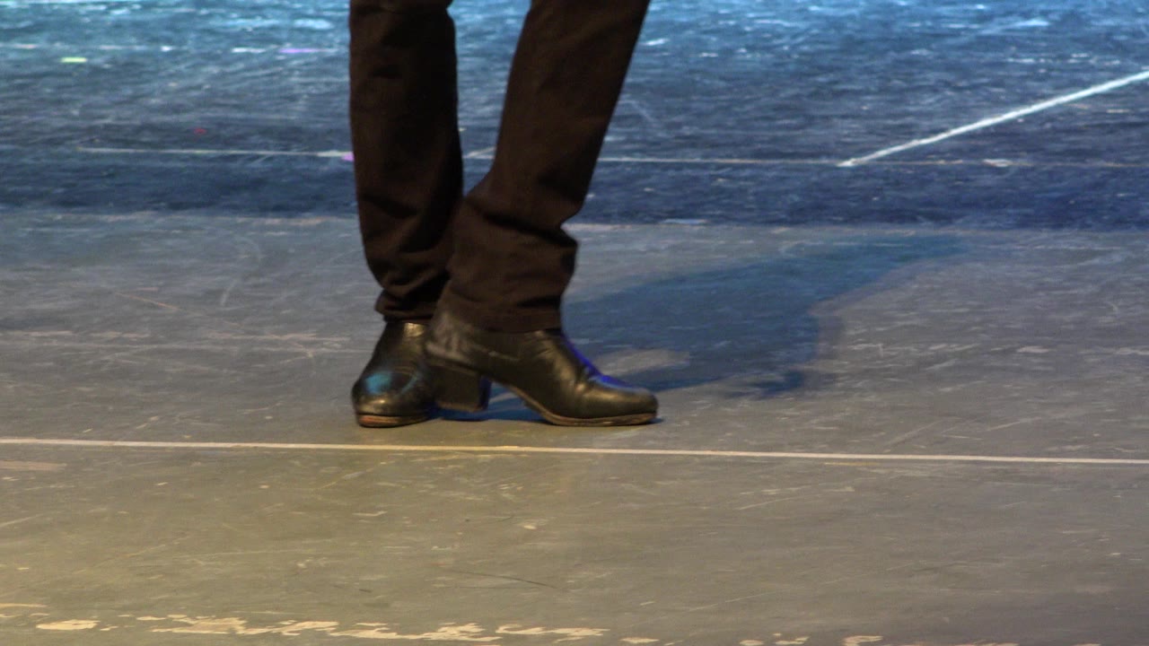 Lower legs in dark pants and black heeled shoes on a scuffed floor.