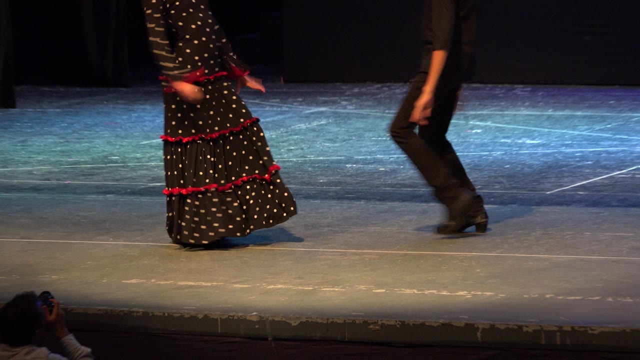 Two teenage dancers are partly cropped as they move across a theater stage in Cabo San Lucas, Mexico, with blue floor lighting and a dark curtain behind them.
