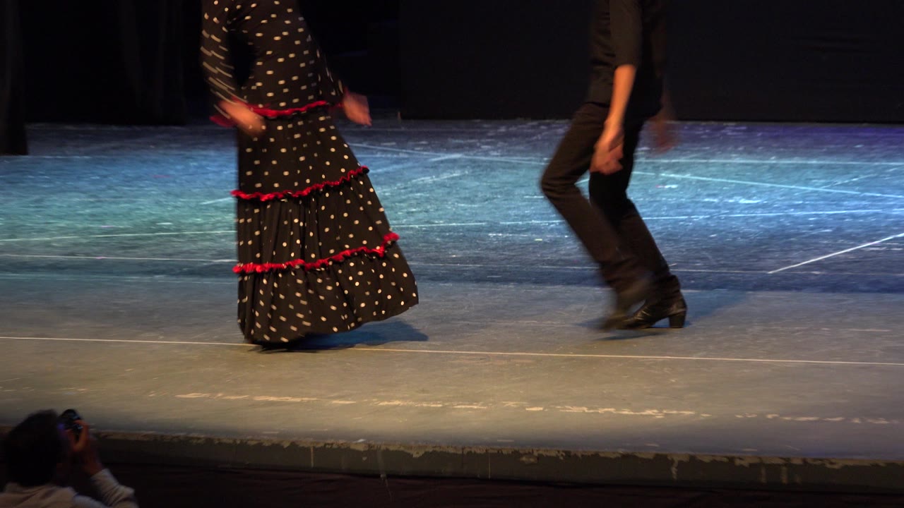 The teenage girl in a dotted black dress and a teenage boy in black are visible mid-step on the Cabo San Lucas theater stage, with their bodies partly cropped by the frame edge.