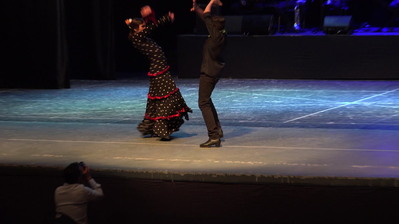 Two dancers raise their arms on a theater stage with musicians in back.