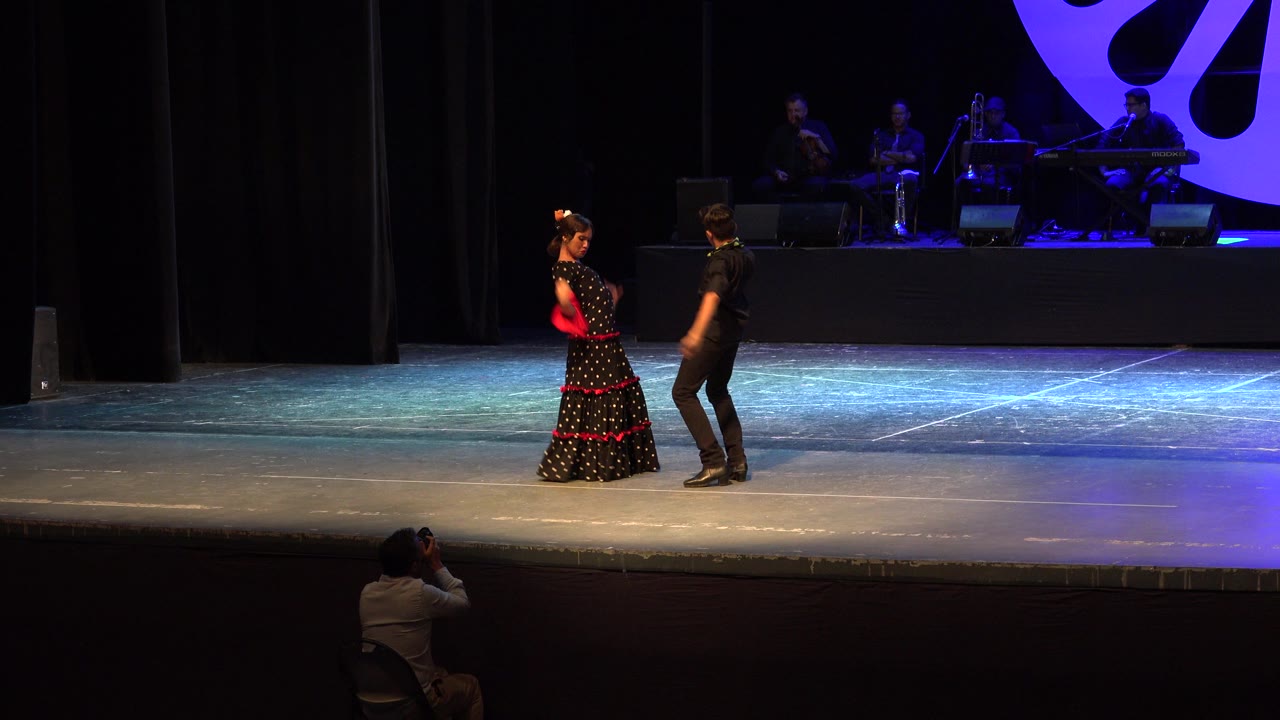 At the start of the Cabo San Lucas clip, a young girl in a black dress with red trim faces a young boy in black on a lit stage while musicians sit behind them.