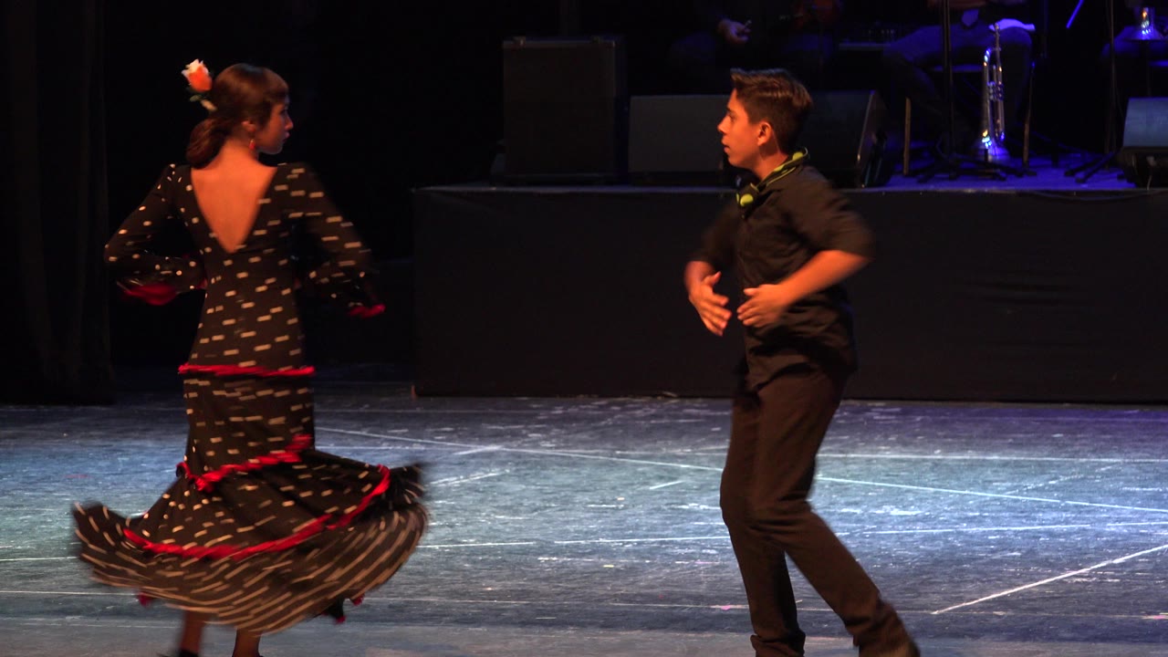 Two young dancers on a dark stage, one turning in a black dress with red trim.