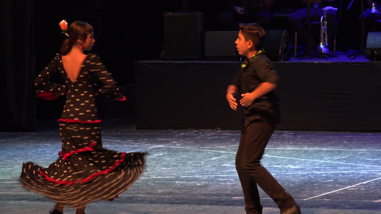 In Cabo San Lucas, Mexico, the girl continues a turn with her skirt lifted by motion while the boy in black steps toward her under blue stage light.