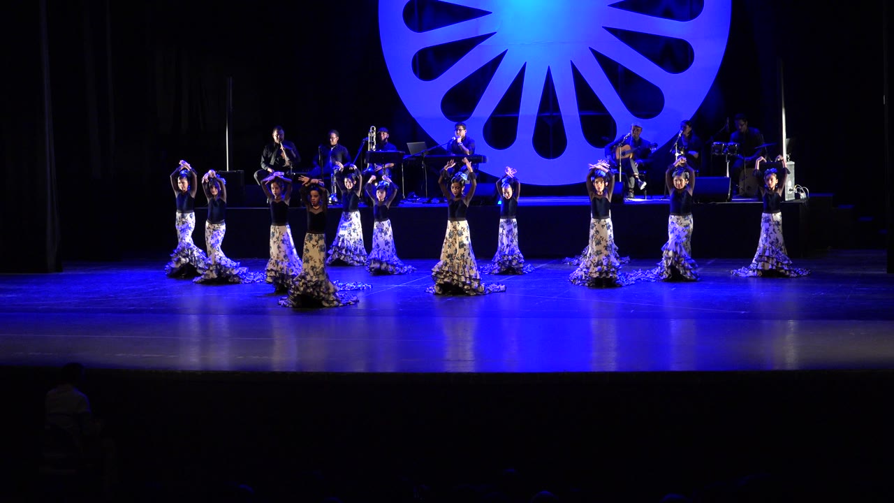 Women dancers in Cabo San Lucas hold raised arm positions across a blue-lit theater stage while musicians remain seated on the platform behind them.