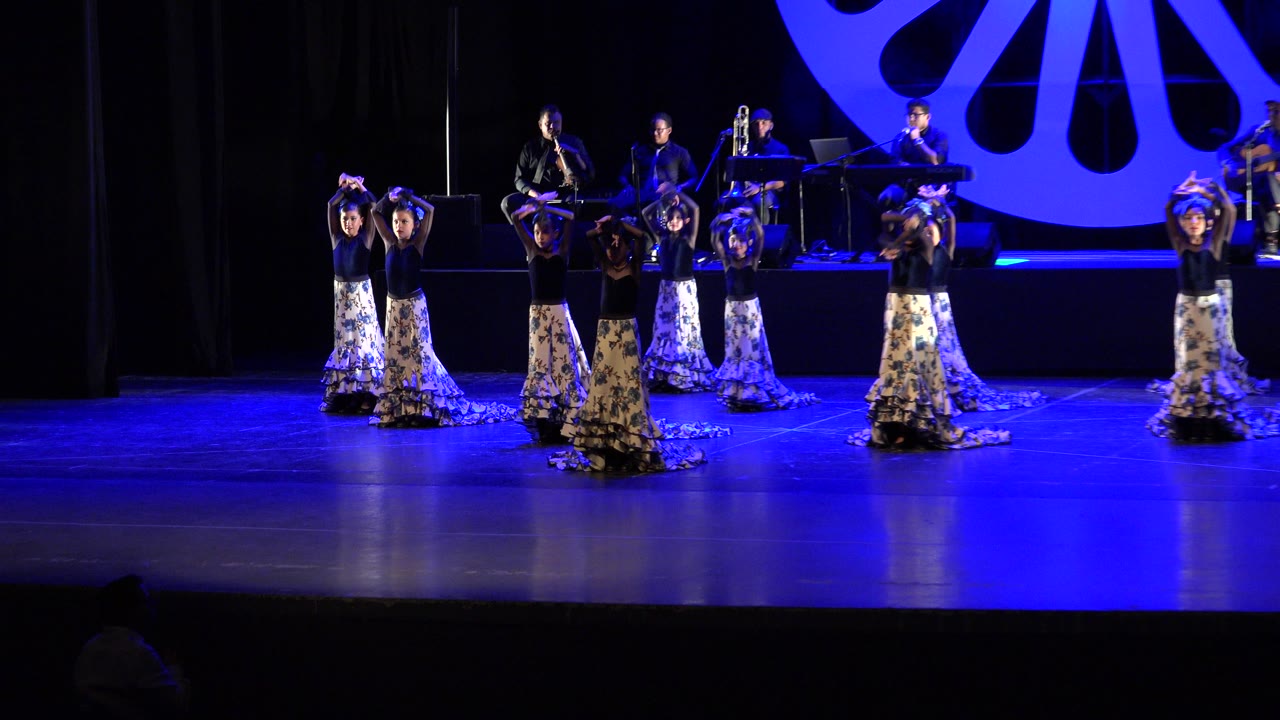 Dancers in long floral skirts performing on a blue-lit stage with musicians behind them.
