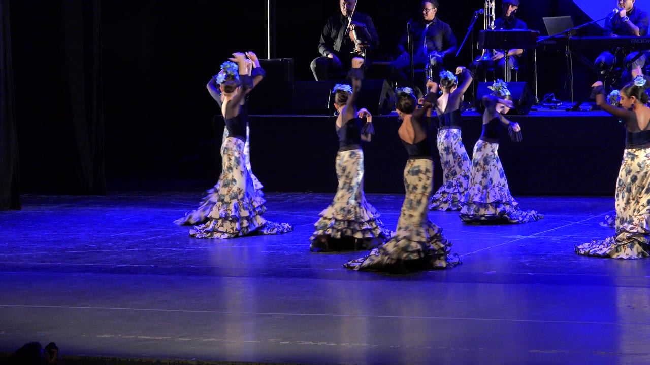 Girls in patterned skirts dance on a blue-lit stage with musicians behind them.