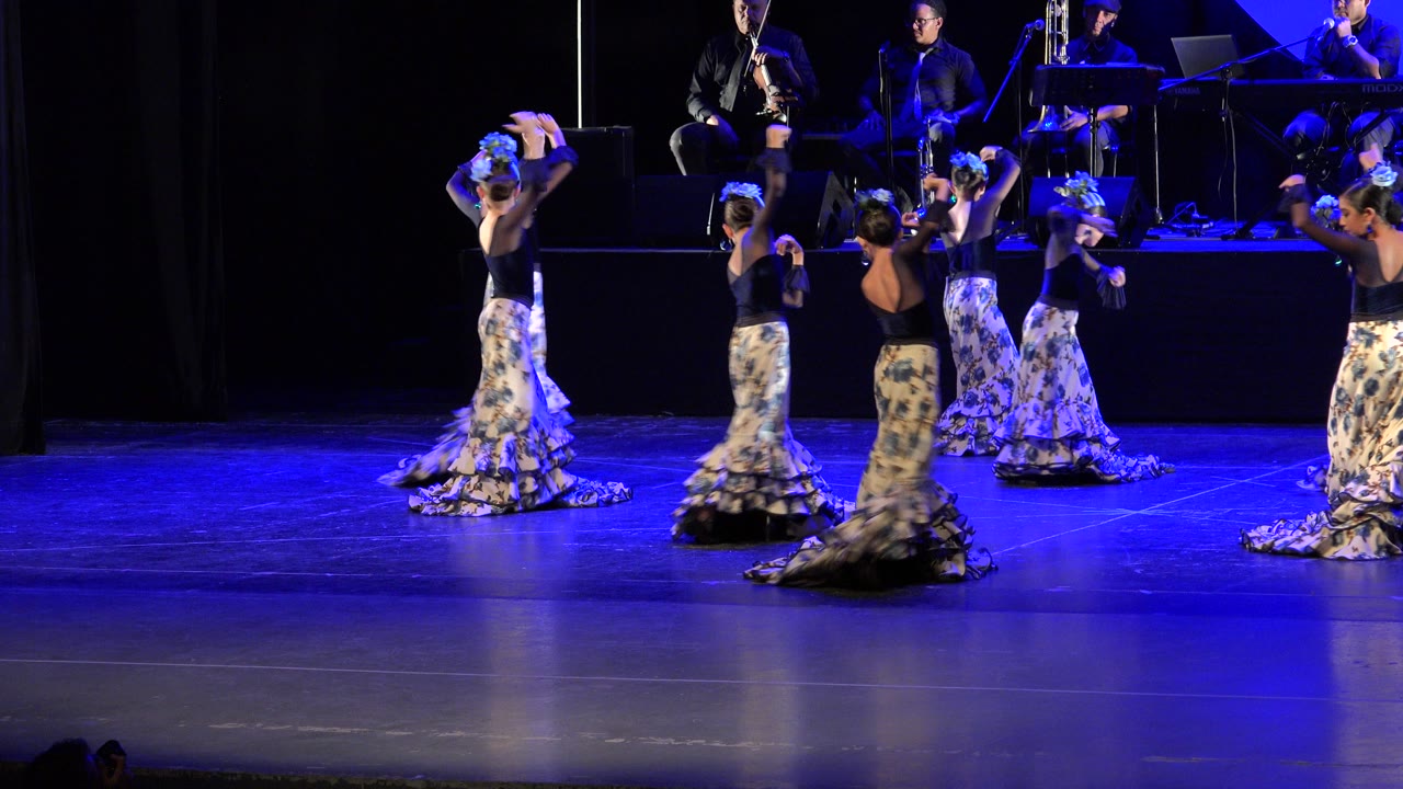 The dancers hold raised-arm poses in Cabo San Lucas, Mexico, while long floral skirts and blue stage light dominate the foreground.