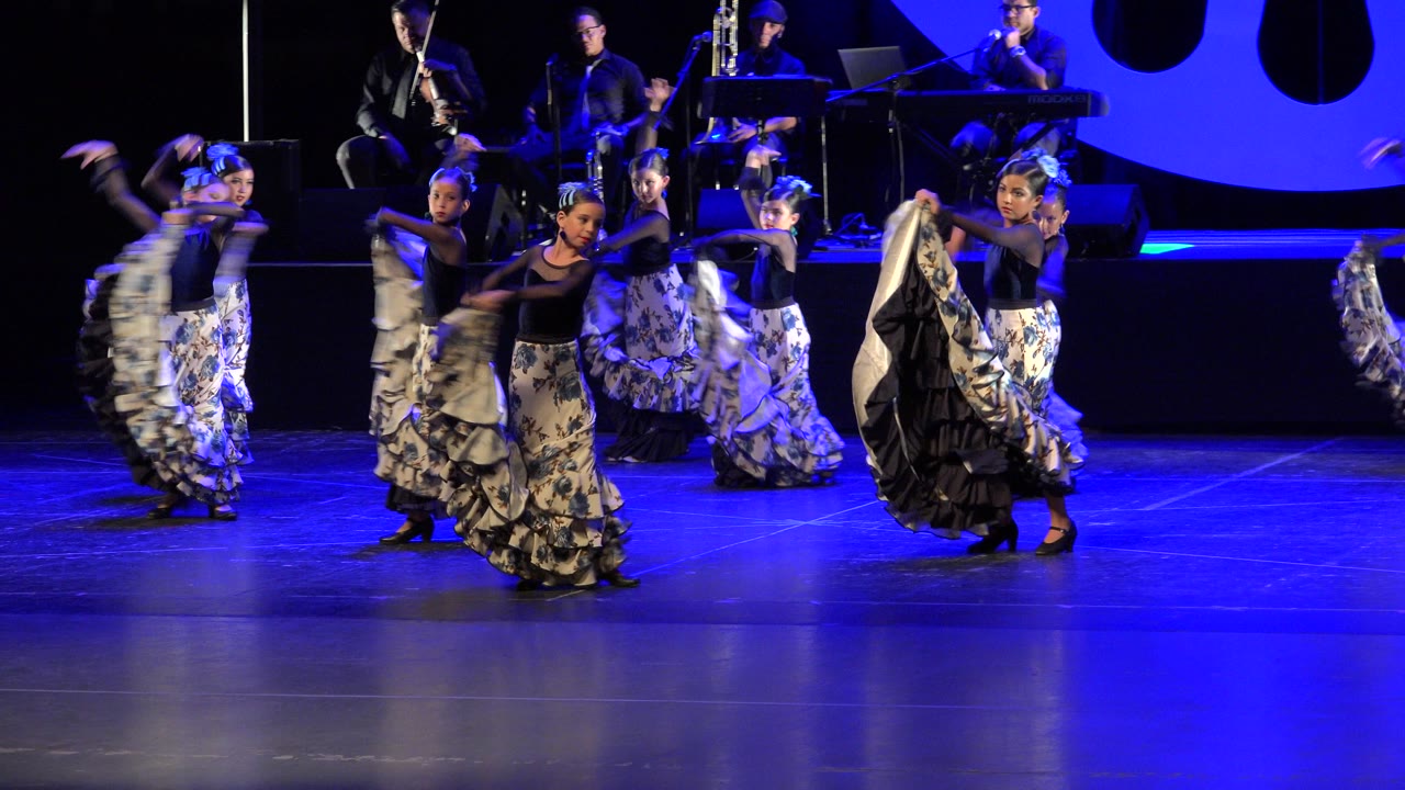 At 0:00 in Cabo San Lucas, Mexico, young girls in patterned ruffled skirts move across a blue-lit stage while seated musicians play behind them.