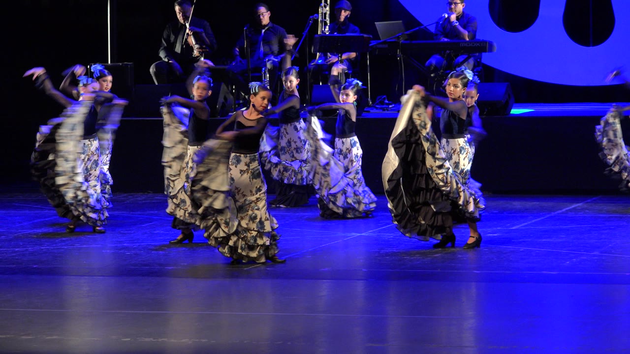 At 0:01, the dancers remain grouped across the stage, with one girl near center holding her skirt forward and musicians seated behind the line.