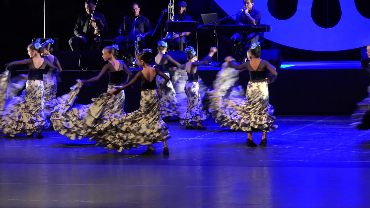 Girls turn across the stage with skirts fanned wide under blue lighting.