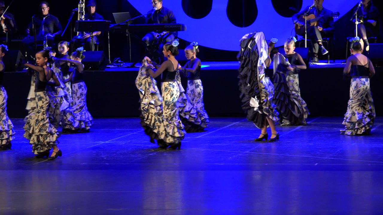 Girls in ruffle skirts dancing on a blue-lit stage with musicians behind them