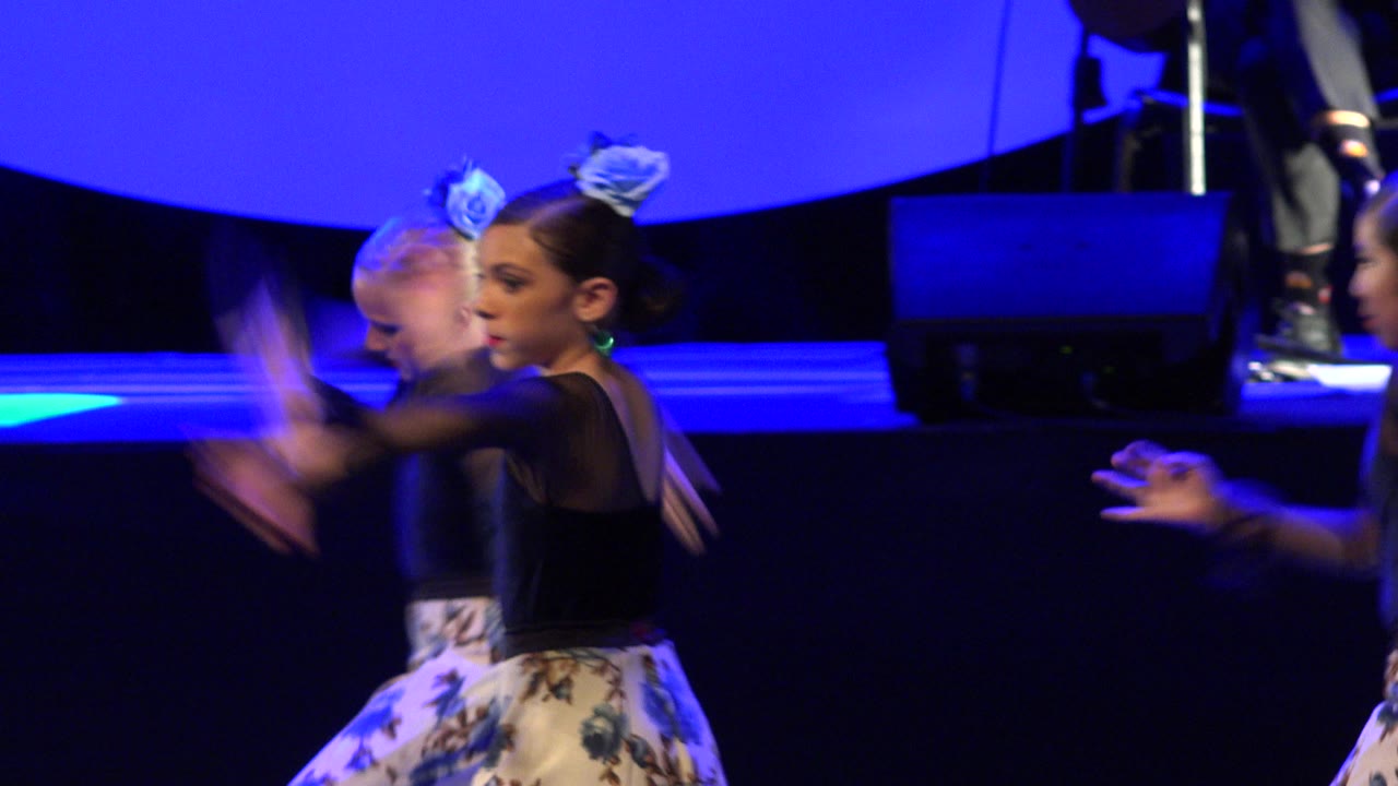 Two young dancers in black tops and floral skirts under blue stage light.