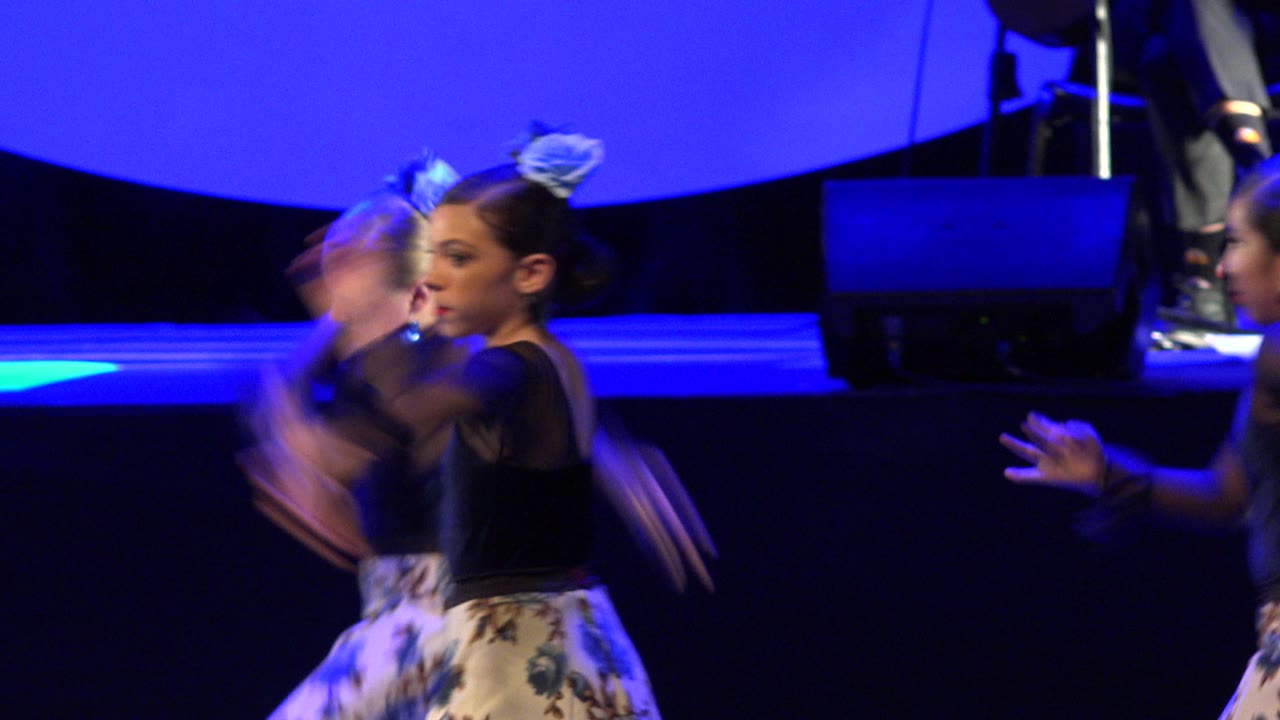 A young dancer in a floral skirt faces right while another child moves past her on the blue-lit performance stage in Cabo San Lucas, Mexico.