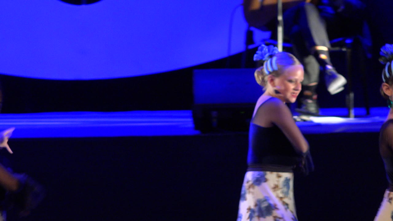 Young dancer on a blue-lit stage with musicians behind her.