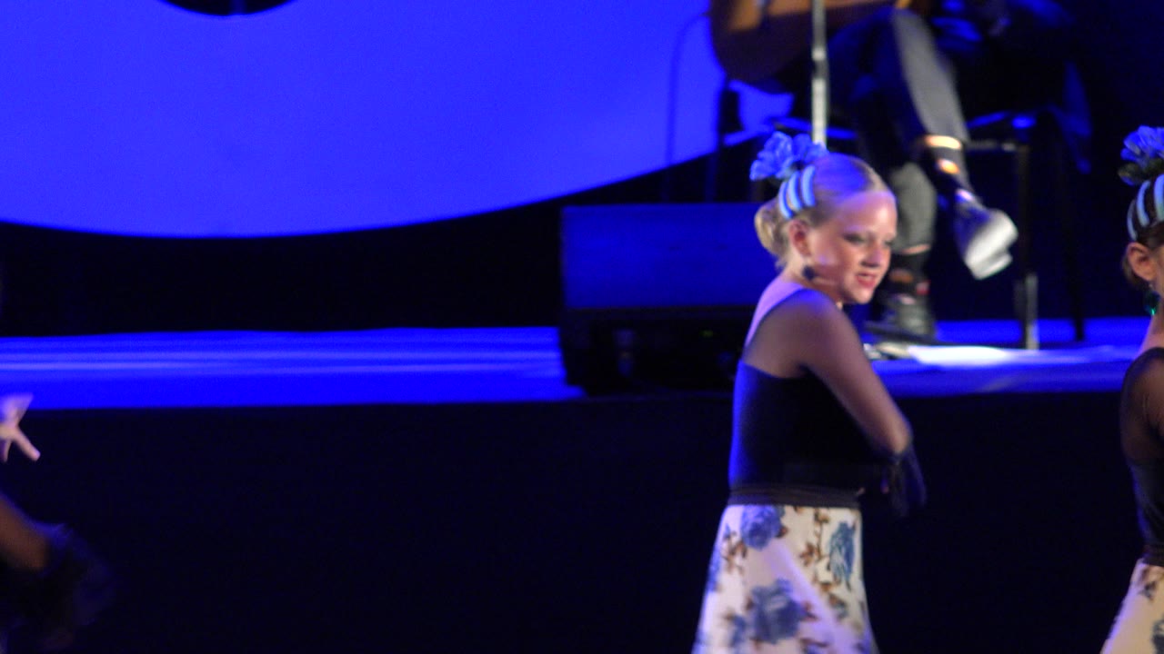 The same young dancer is framed near center on the Cabo San Lucas stage, looking sideways while a dark stage monitor and musician legs sit in the blue-lit background.