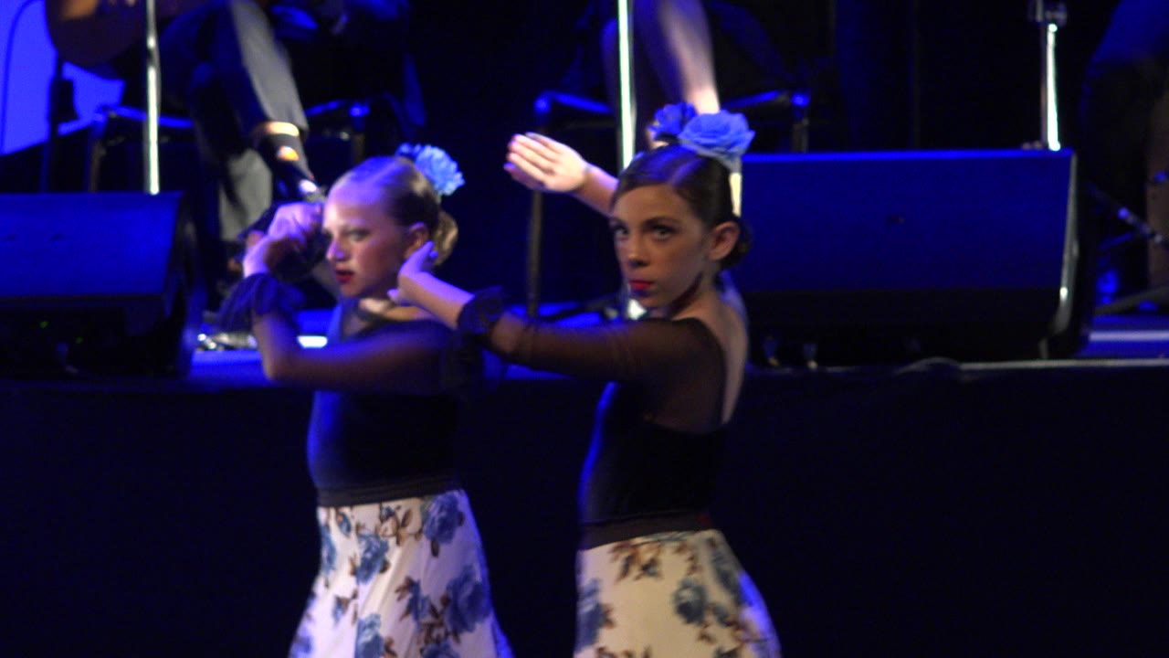 The two young dancers hold a similar forward-facing pose in Cabo San Lucas, Mexico, with one girl's arms raised and blue stage light falling across their costumes.