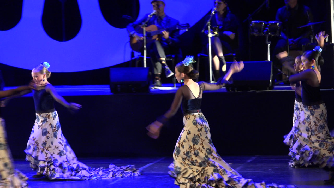 Young girls in floral skirts dancing on a blue-lit stage with musicians behind them.