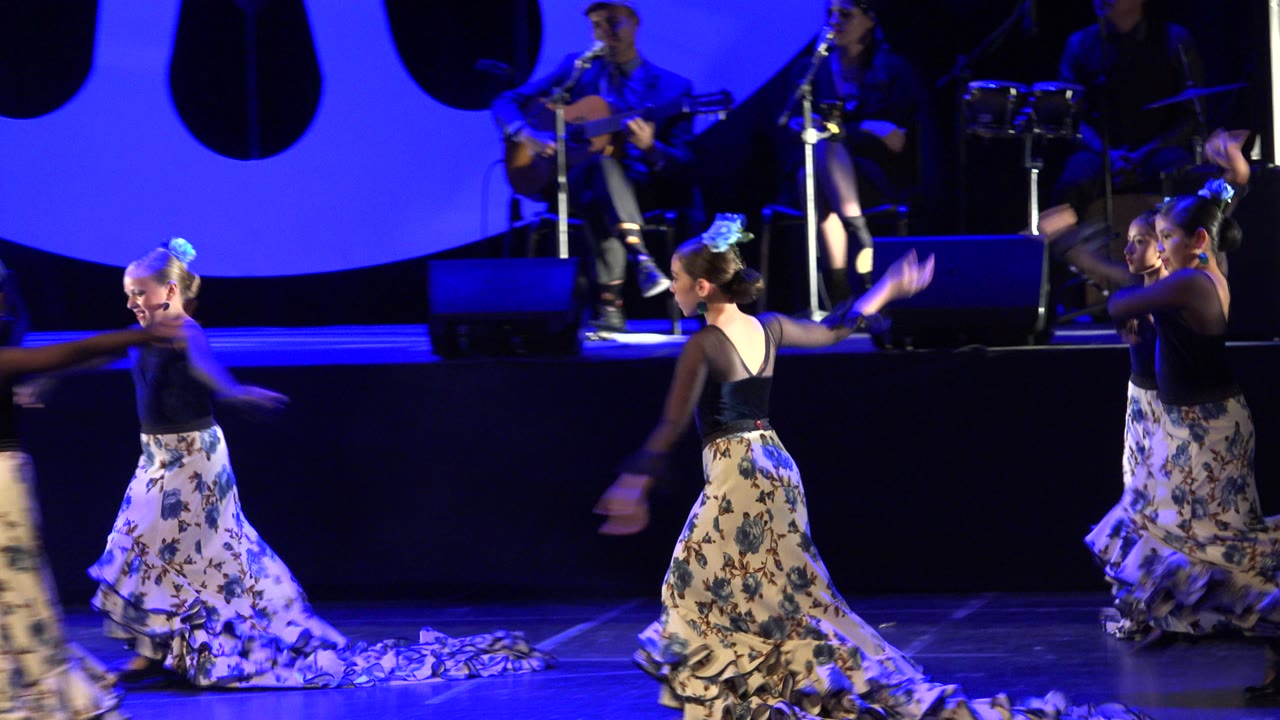 A wide stage view in Cabo San Lucas, Mexico shows child dancers shifting positions, including one turned away from camera, while musicians sit behind them.