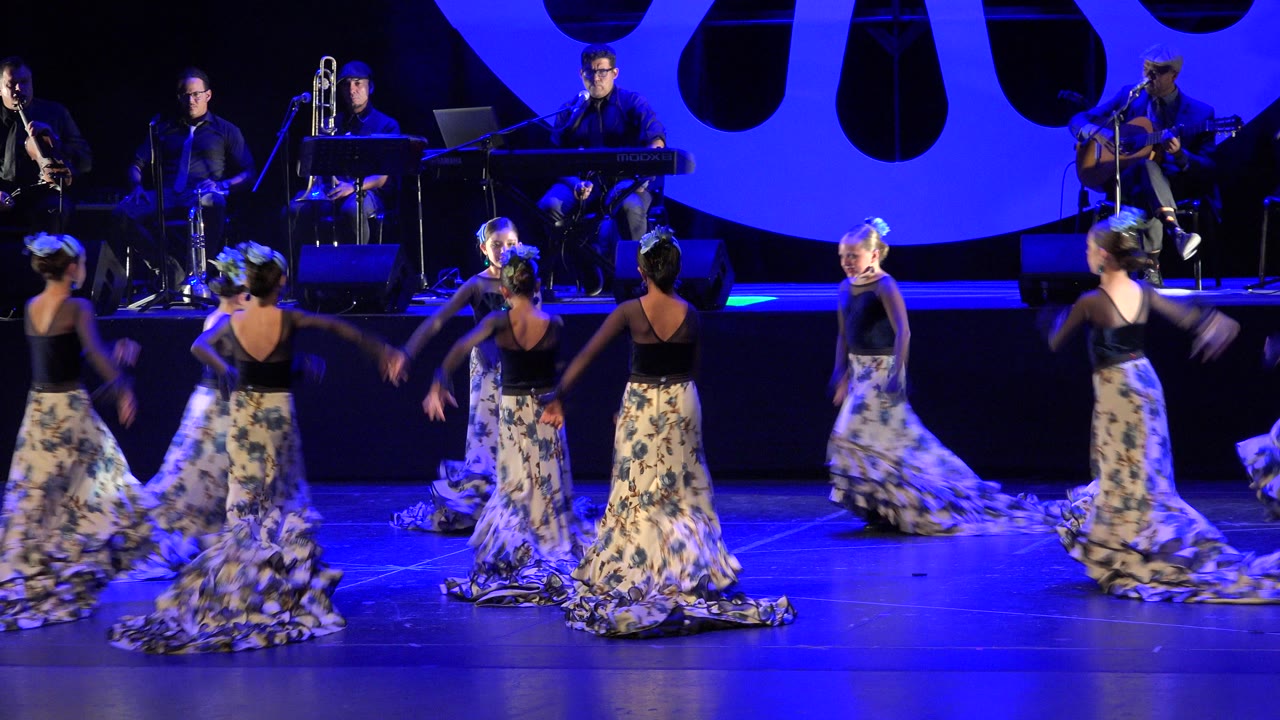 Dancers in patterned skirts perform on a blue-lit stage with musicians behind them.