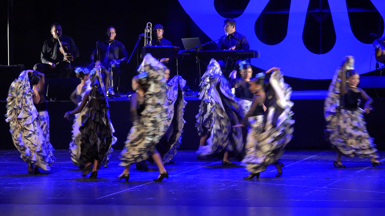 Young dancers cross a blue-lit stage in front of seated musicians.