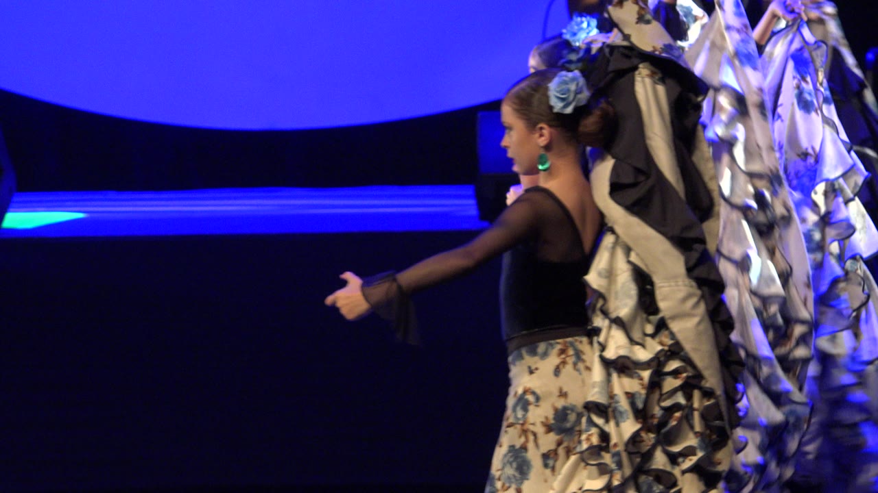 At 0:01 in Cabo San Lucas, Mexico, the same child dancer extends her arm while other girls in ruffled patterned costumes cluster behind her on the blue-lit stage.