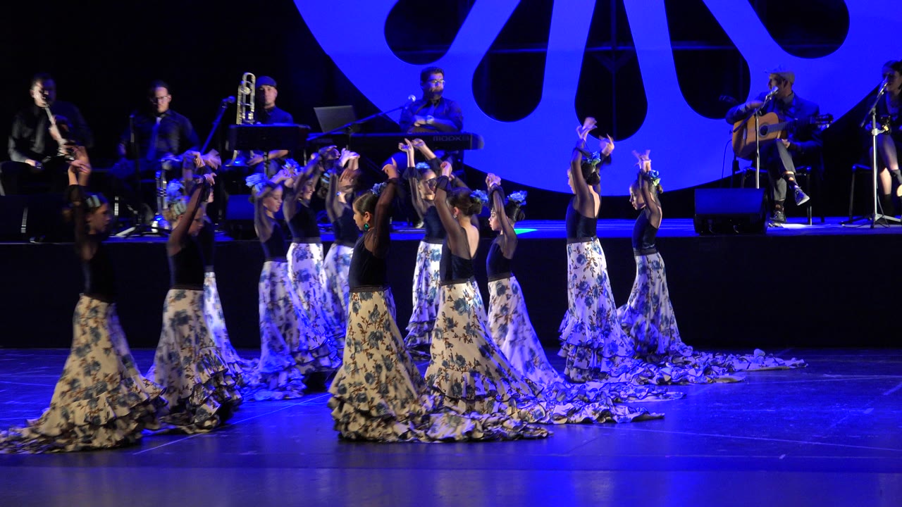 The dancers cluster near center stage in Cabo San Lucas, Mexico, with arms lifted and long patterned skirts trailing under blue theatrical light.