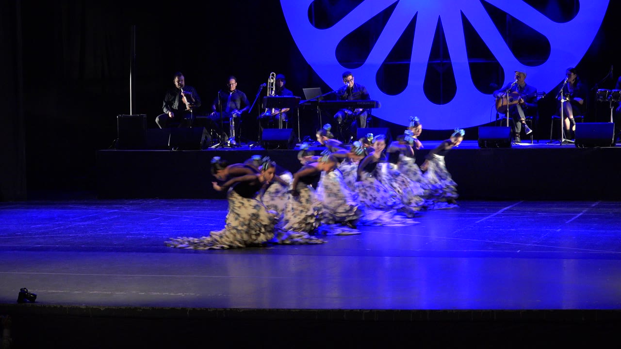 At the start of the Cabo San Lucas performance, a line of girls in black tops and patterned skirts kneels low across the foreground while musicians sit behind them.