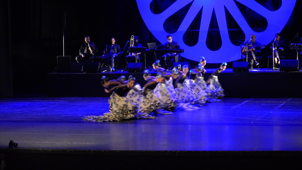 Girls remain low to the floor during the Cabo San Lucas stage routine as the live band and large blue fan-shaped set piece fill the background.